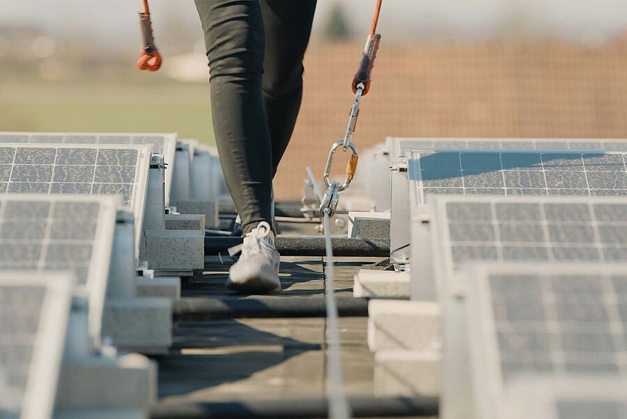 Een persoon beweegt zich op een plat dak tussen fotovoltaïsche panelen, beveiligd door individuele bescherming met PBM's en een touwsysteem.